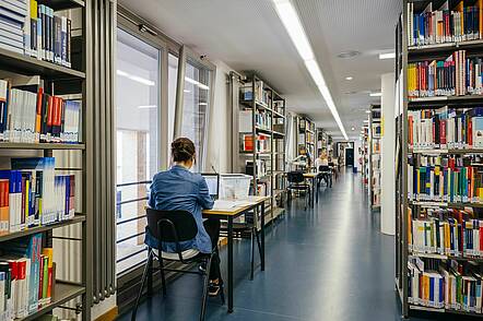 Foto: zwei Personen sitzen an Einzeltischen an einem langen Gang in einer Bibliothek 