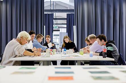Foto: Studierende sitzen zusammen an einem großen Tisch und üben sich in Malereitechniken.