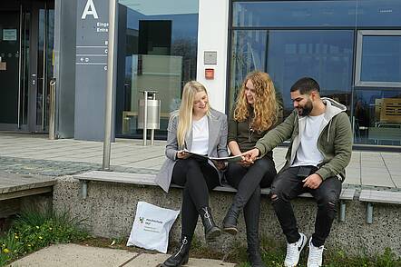 Foto: Studierende unterhalten sich sitzend vor dem Eingang zur Hochschule Hof.