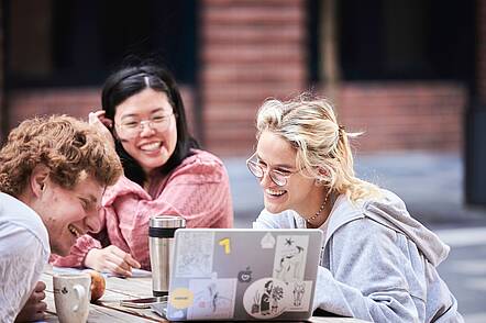 Foto: Zwei Studentinnen und ein Student sitzen an einem Tisch auf dem Campus der Hochschule Fulda und blicken in einen Laptop