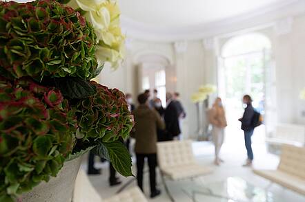 Foto: Blick auf eine Gruppe von Personen, die in einem eleganten Raum zusammenstehen und im Vordergrund stehen Blumen in einer Vase