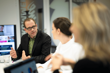 Foto: Ein Mann und eine Frau unterhalten sich am Arbeitsplatz vor Bildschirmen.