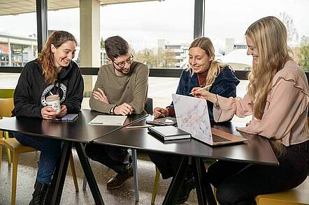 Foto: Studierende sitzen an einem Tisch mit Laptop und unterhalten sich