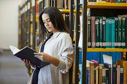 Foto: Eine Studierende der RPTU Kaiserslautern lehnt an einem Regal in der Bibliothek und blickt in ein Buch.