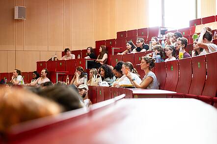 Foto: Studierende sitzen in einem holzvertäfelten Hörsaal auf roten Bänken