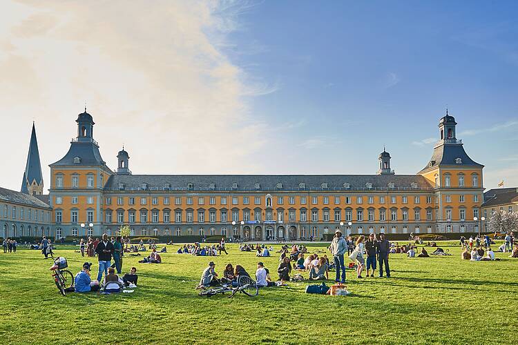 Foto: Studierende sitzen auf der Hofgartenwiese vor dem Hauptgebäude der Universität Bonn.