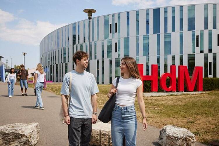 Foto: Studierende gehen über den Campus der Hochschule der Medien Stuttgart mit der Hochschule und dem Logo im Hintergrund