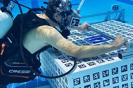 Foto: Ein Taucher in einem Schwimmbecken trägt ein VR-Headset und präsentiert eine neue VR-Anwendung. Vor ihm liegt ein Block mit Tracking-Code. 