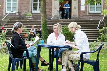 Foto: Drei Personen sitzen an einem Tisch auf dem Campus vor der Fliedner Fachhochschule Düsseldorf und unterhalten sich.