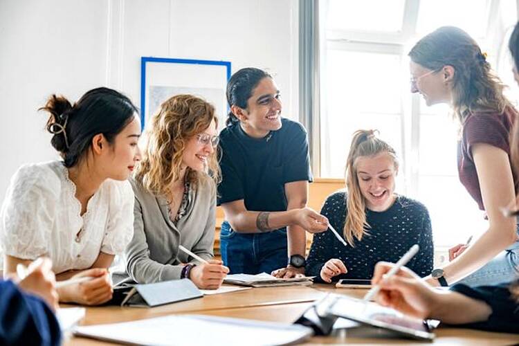 Foto: Eine Gruppe Studierender sitzt in einem Raum, diskutiert und macht sich Notizen.