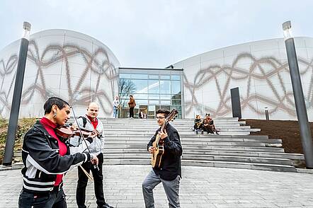 Foto: Studierende stehen vor dem Institut für Musik der Hochschule Osnabrück und musizieren mit Geige und Gitarre