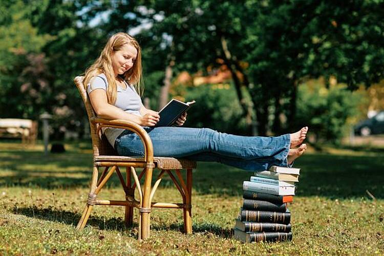 Foto: Eine Studentin sitzt auf einem Stuhl auf einer Wiese, hat die Beine auf einen Stapel Bücher gelegt und liest ein Buch