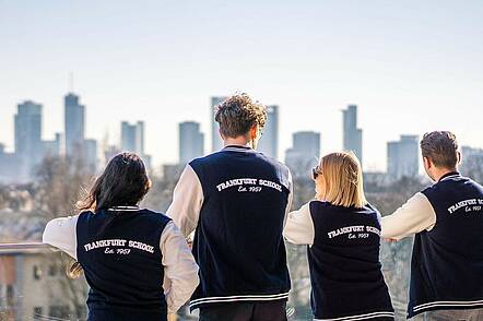 Foto: Blick auf vier Studierende, die alle eine blau-weiße Universitätsjacke tragen, mit der Skyline von Frankfurt im Hintergrund 