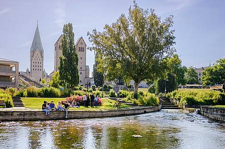 Foto: Studierende der Universität Paderborn sitzen auf einer Wiese an einem kleinen See und unterhalten sich