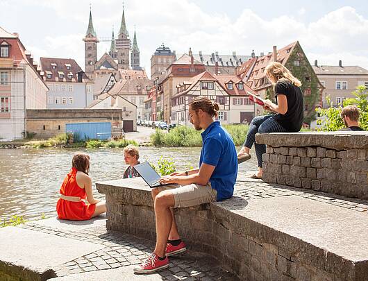 Foto: Studierende sitzen am Ufer des Mains, im Hintergrund Bamberg