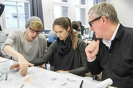 Foto: Zwei Studierende der Architektur sitzen mit Ihrem Dozenten am Tisch vor einem Plan