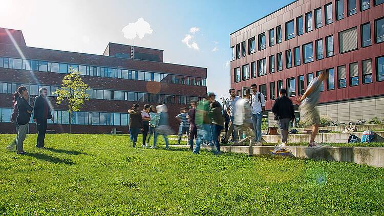 Foto: Auf einem Rasen vor zwei Gebäuden stehen Studierende in Gruppen und unterhalten sich