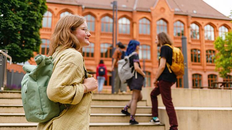 Foto: Eine Studentin geht über den Campus der HAWK, vorbei an Treppen und entlang eines Backsteinhochschulgebäudes