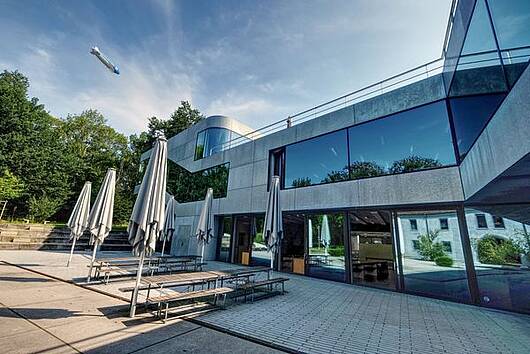 Foto: Blick auf den Außenbereich der Cafeteria der Zeppelin Universität mit Tischen, Bänken und Sonnenschirmen
