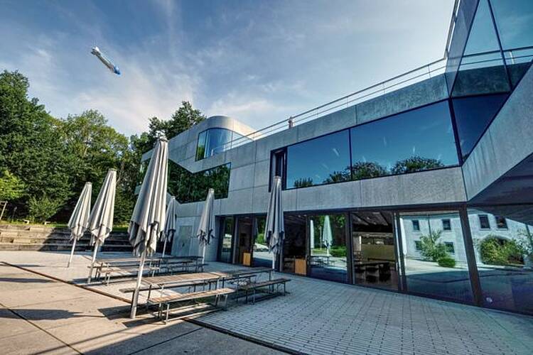 Foto: Blick auf den Außenbereich der Cafeteria der Zeppelin Universität mit Tischen, Bänken und Sonnenschirmen