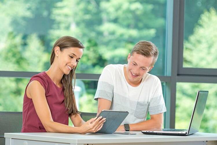 Foto: Zwei Studierende sitzen an einem Tische vor einem Notebook und einem Tablet.