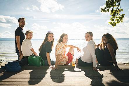 Foto: Studentinnen und Studenten sitzen auf einem Steg am See und blicken in die Kamera des Fotografen