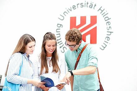 Foto: Studierende stehen vor dem Logo der Universität Hildeheim und blicken in ein Buch.