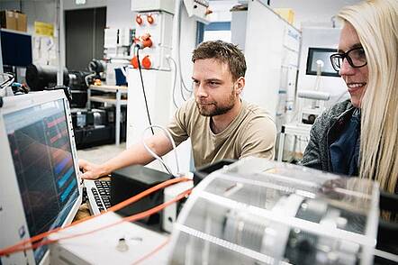 Foto: Zwei Studierende sitzen an einem Tisch in einem Labor vor einem technischen Experiment.