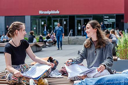 Foto: Zwei Studentinnen sitzen auf dem Campus vor dem Hörsaalgebäude und lernen zusammen.