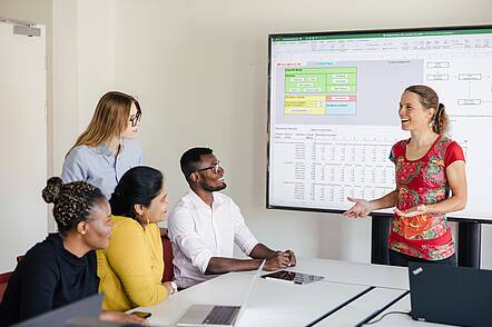Foto: Vier Studierende sitzen in einem Seminarraum und folgen den Ausführungen der Dozentin, hinter ihr ein großer Bildschirm mit Tabellen 