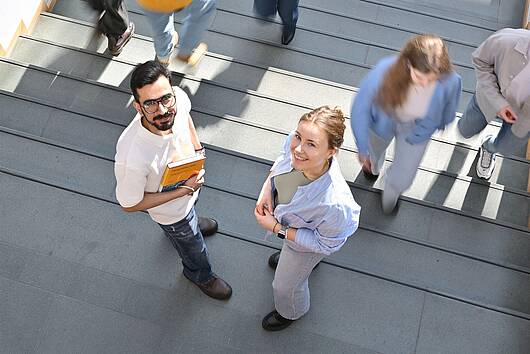 Foto: Zwei Studierende stehen auf einer Treppe, halten Bücher in der Hand und blicken in die Kamera