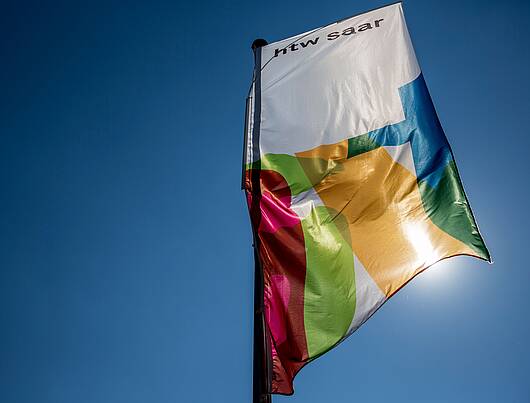 Foto: Das Logo der Hochschule für Technik und Wirtschaft des Saarlandes auf eine Fahne im Wind