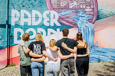 Foto: Eine Gruppe Studierender steht vor einer Wand und betrachtet ein großes Grafiti der Universität Paderborn