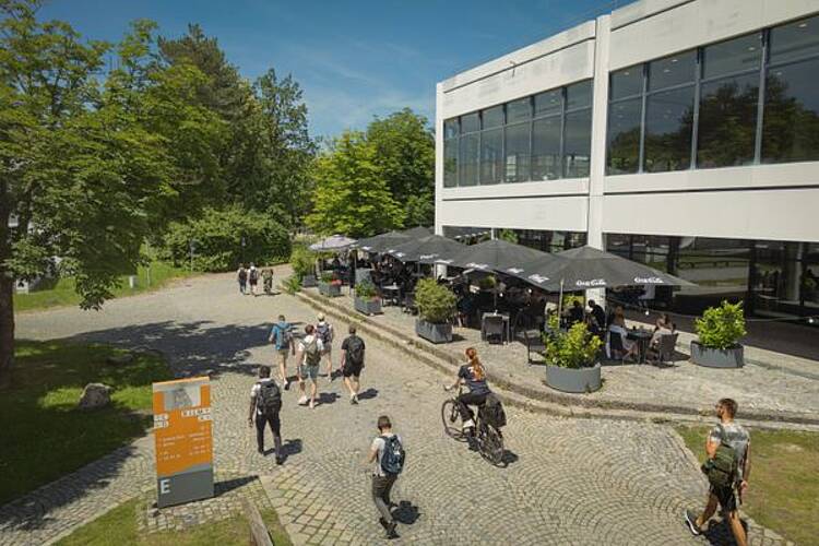 Foto: Studierende gehen oder fahren mit dem Fahrrad über den Campus, im Hintergrund Sonnenschirme und Menschen, die darunter sitzen 