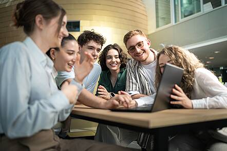 Foto: Sechs Studierende sitzen an einem Tisch und blicken gemeinsam in einen Laptop