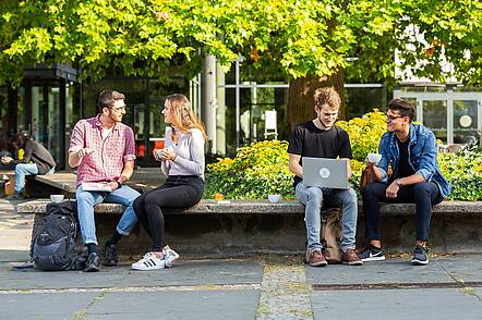 Foto: Vier Studierende sitzen auf einer Steinbank auf dem Campus der TU Berlin und unterhalten sich