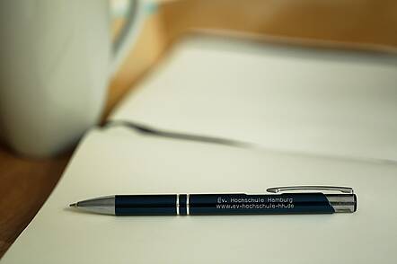 Foto: Ein Kugelschreiber der Evangelischen Hochschule für Soziale Arbeit & Diakonie liegt auf einem Blatt Papier und daneben steht eine weiße Tasse