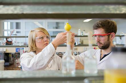 Foto: Zwei Studierende mit Schutzbrillen in einem Labor, im Vordergrund ein Regal mit Reagenzgläsern