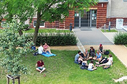 Foto: Eine Gruppe Studierender sitzt auf einer Wiese auf dem Campus der Katholischen Hochschule für Sozialwesen Berlin und unterhält sich.