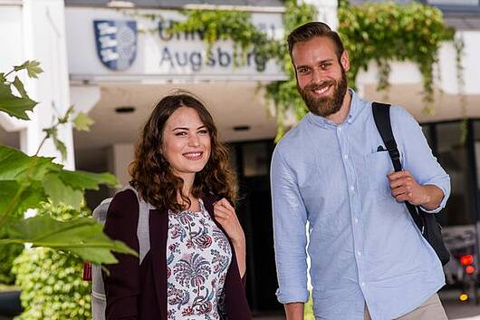 Foto: Zwei Studierende der Universität Augsburg stehen vor dem Eingangsportal der Universität