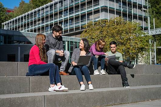 Foto: Eine Gruppe Studierender sitzt auf einer Treppe vor dem Campusgebäude der Universität des Saarlandes
