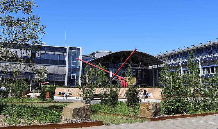 Foto: Blick über den Campus der Hochschule Bonn-Rhein-Sieg mit Studierenden, die auf einer Holzbank vor einer roten Skulptur sitzen