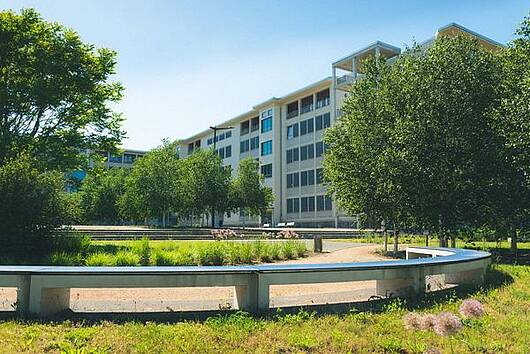 Foto: Blick auf den Campus der Hochschule Merseburg mit einer runden Sitzbank und Bäumen im Vordergrund