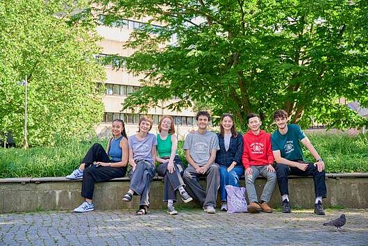 Foto: Studierende der Universität Hamburg  sitzen gemeinsam auf einer Bank auf dem Campus.