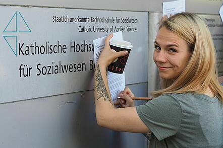 Foto: Eine Studentin steht vor dem Logo der KHS mit einem Becher Kaffee und Schreibunterlagen.