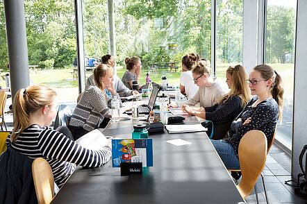 Foto: Studierende sitzen an einem langen Tisch in der Mensa und lesen oder unterhalten sich