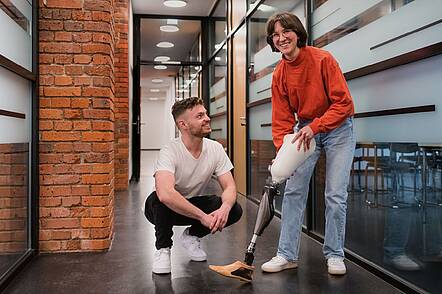 Foto: Zwei Studierende testen eine Beinprothese, einer kniet dabei, die andere steht und hält die Prothese