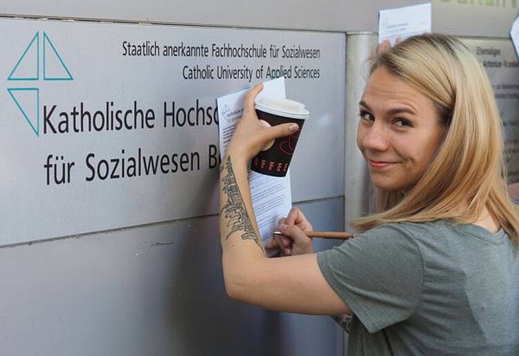 Studentin der Katholischen Hochschule für Sozialwesen Berlin (Foto: Katholische Hochschule für Sozialwesen Berlin) Foto: Eine Studentin steht vor dem Logo der KHS mit einem Becher Kaffee und Schreibunterlagen.