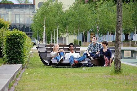 Foto: Studierende sitzen auf einer Bank auf dem Campus und unterhalten sich.