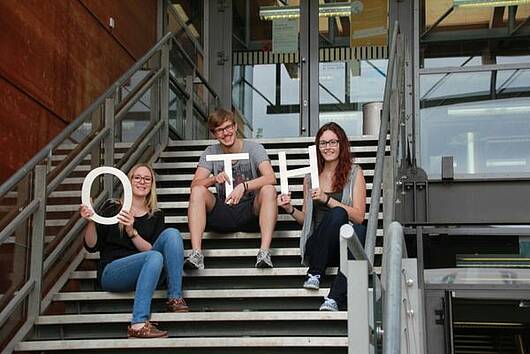 Foto: Gruppe Studierender sitzt auf einer Treppe vor einem Gebäude und hält die Buchstaben OTH hoch